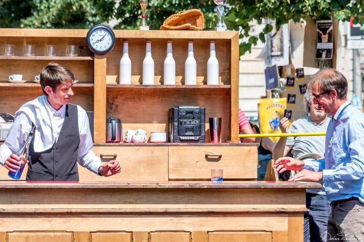 Deux comédiens devant un bar, un serveur tenant une bouteille et l'autre au bout du bar souriant